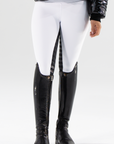 A person wearing white competition tights with a black seat, paired with a black jacket and black riding boots. The tights have silver logo detailing on the back pocket.